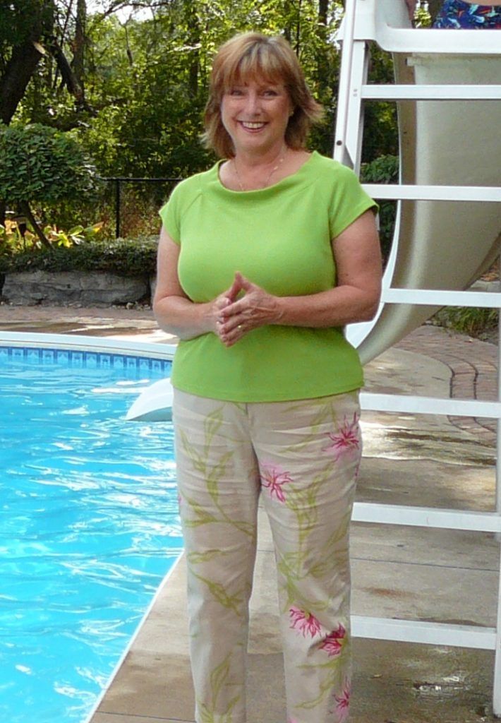 Woman by pool in green top and floral pants. Sunlight, trees, and white pool ladder visible.