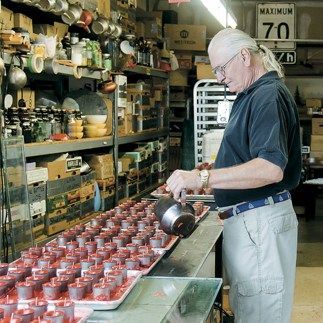 Man pouring liquid from a pot into small containers on a tray in a cluttered workshop.