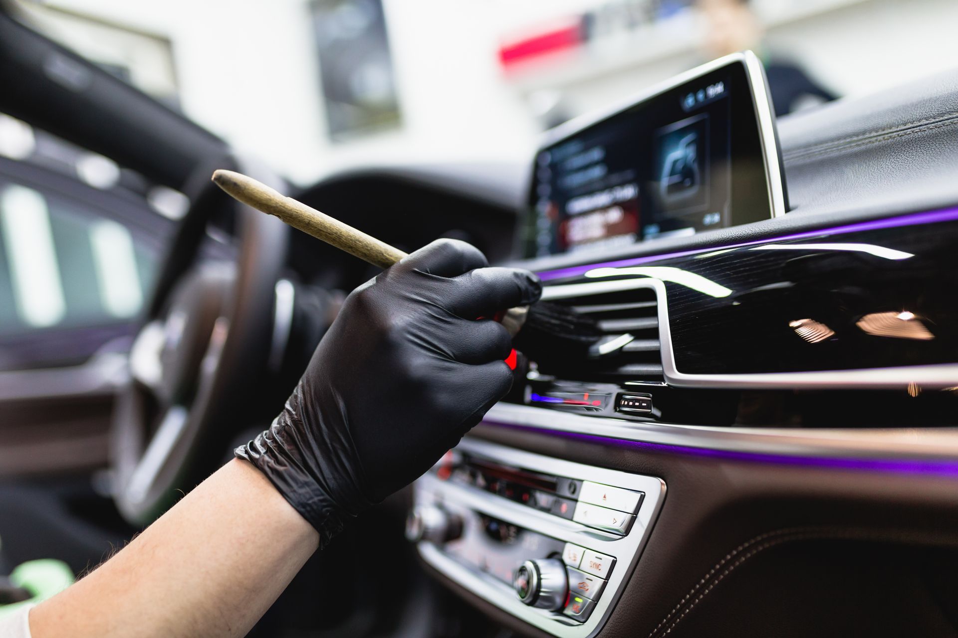 Gloved hand cleaning a car dashboard with a brush, showing a touchscreen and center controls