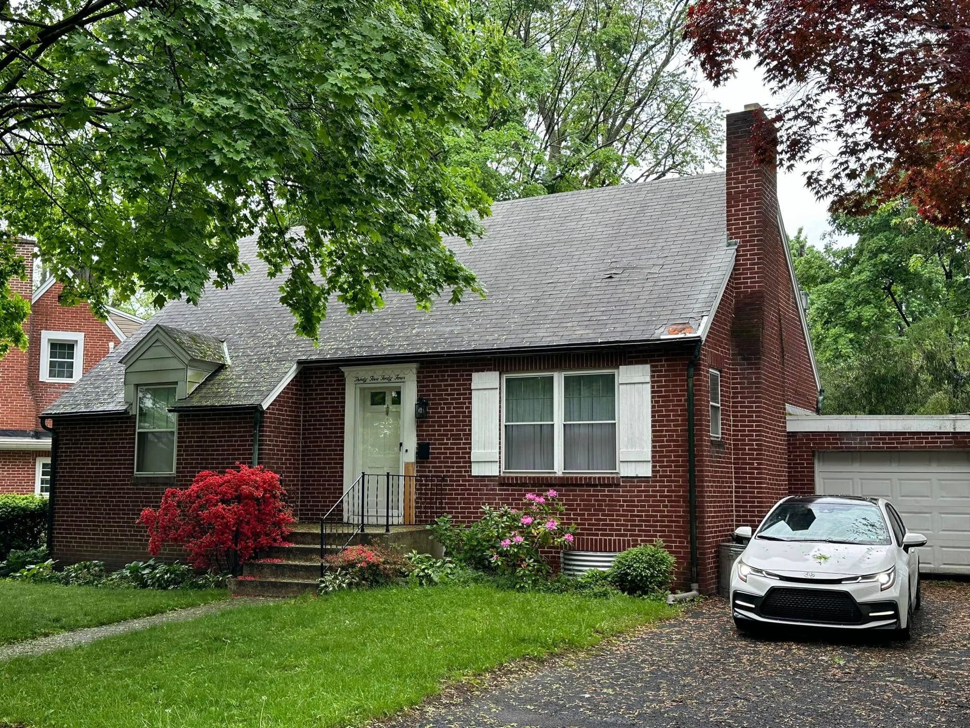 Red brick house with gray roof, green lawn, and a white car parked in the driveway under trees.
