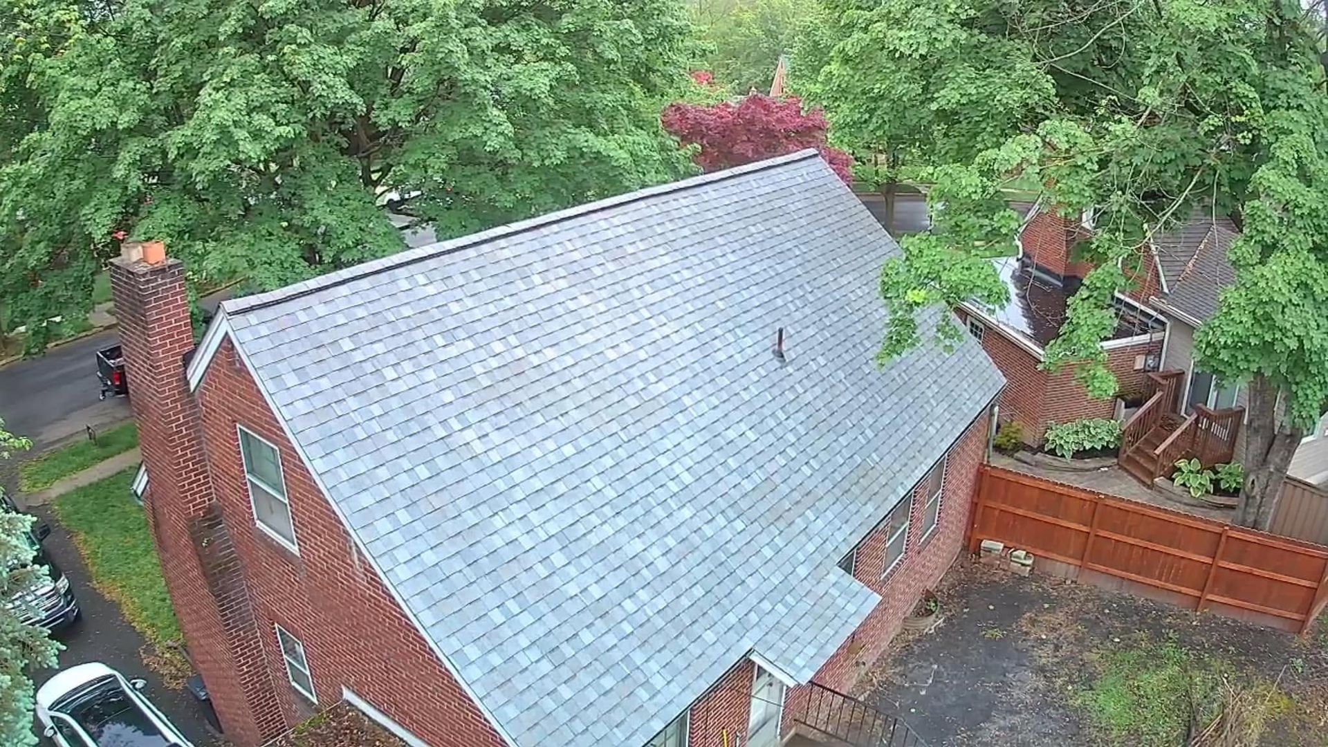 Aerial view of a brick house with a gray roof, surrounded by trees and a fenced yard.