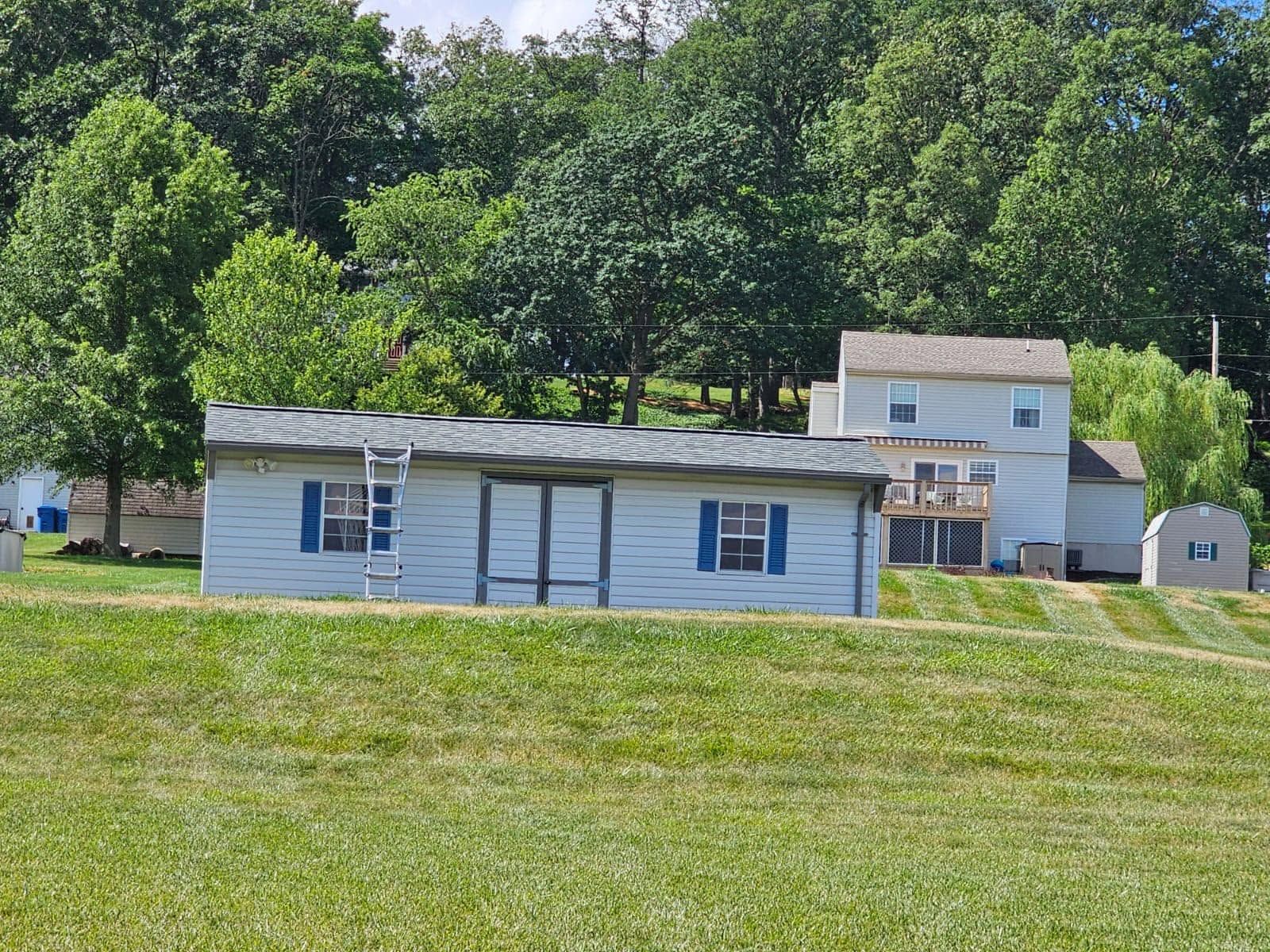 Two-story white house with blue shutters and a grassy lawn, backed by trees on a hillside