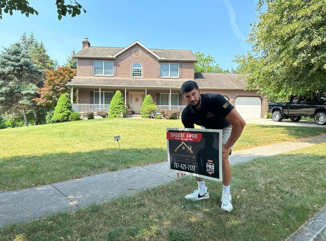 Man carrying a boxed grill on a driveway in front of a brick house, smiling.