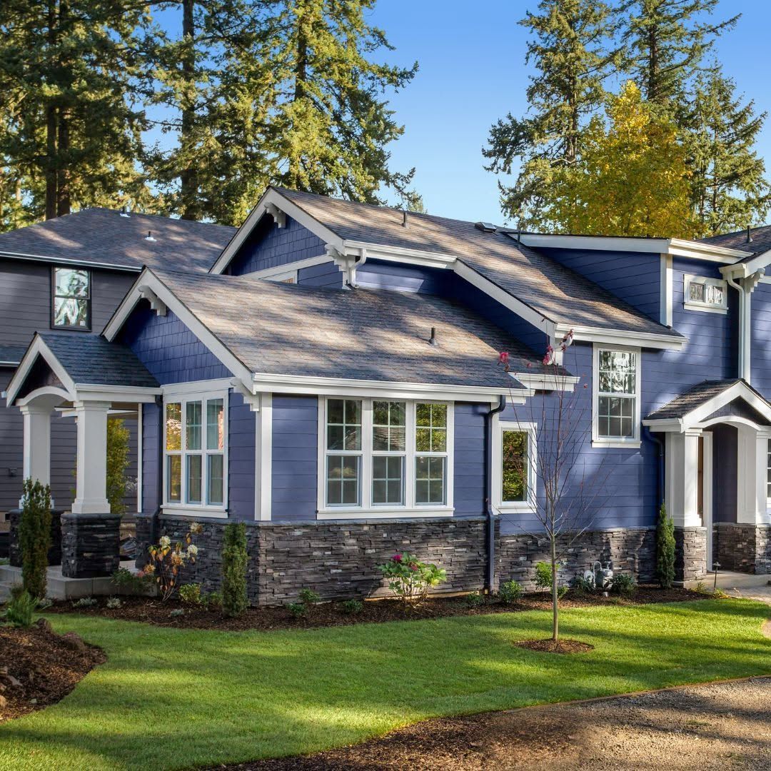 Blue suburban house with stone accents, white trim, and a green lawn surrounded by tall trees.