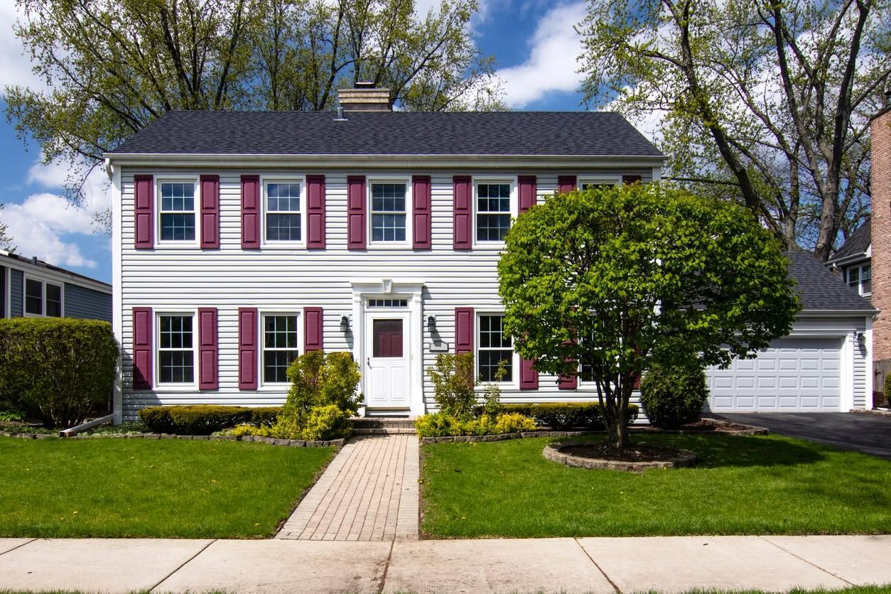 Two-story white house with maroon shutters, front lawn, and a brick walkway under a blue sky