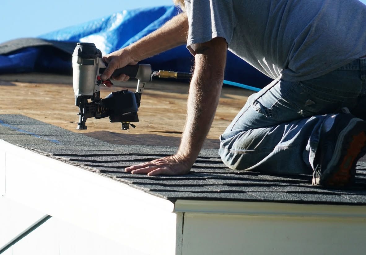 Worker kneeling on a roof, fastening shingles with a nail gun beside a blue tarp