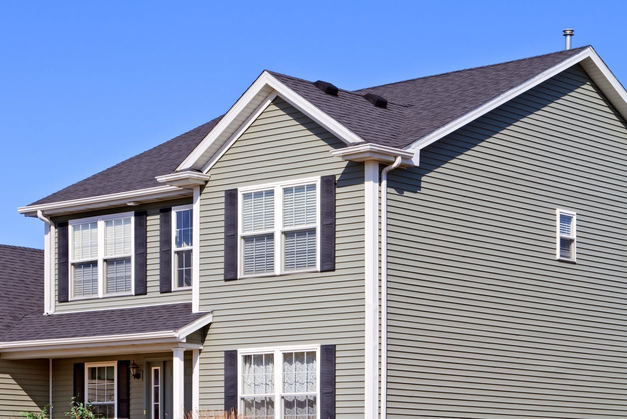Two-story gray house with white trim and a brown shingled roof under a clear blue sky
