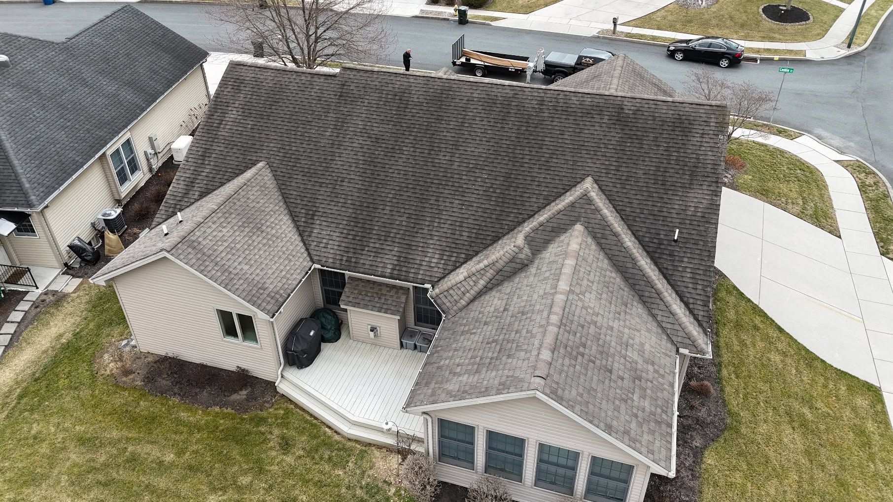 Aerial view of a suburban house with dark shingle roof, attached garage, and driveway on a corner lot.
