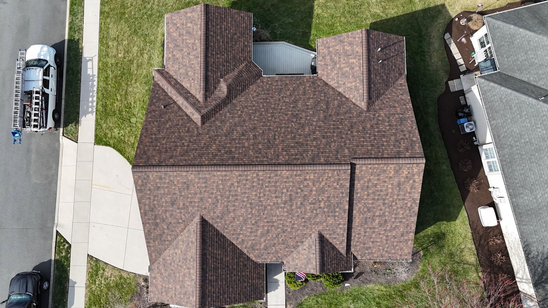 Aerial view of a brown-roofed house with a driveway, sidewalk, and parked vehicle beside a street.