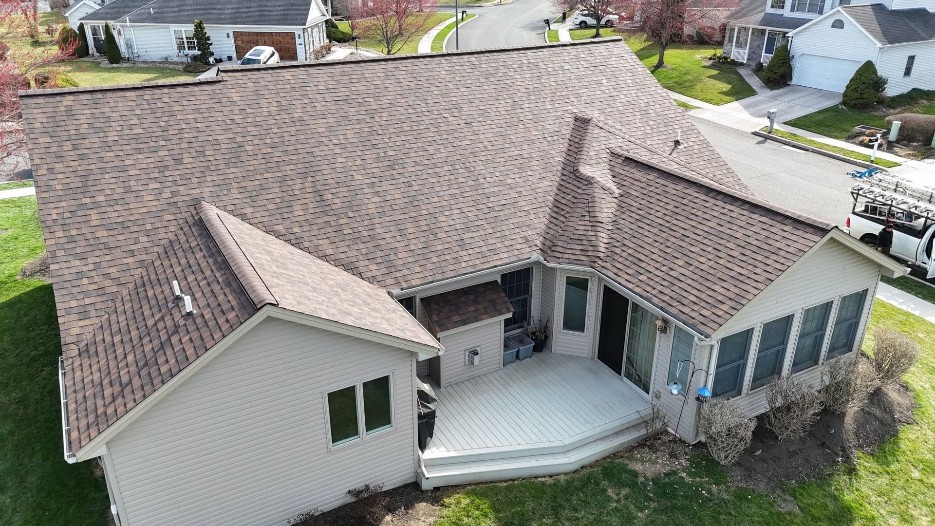 Aerial view of a beige house with a brown shingled roof and covered front porch in a suburban neighborhood