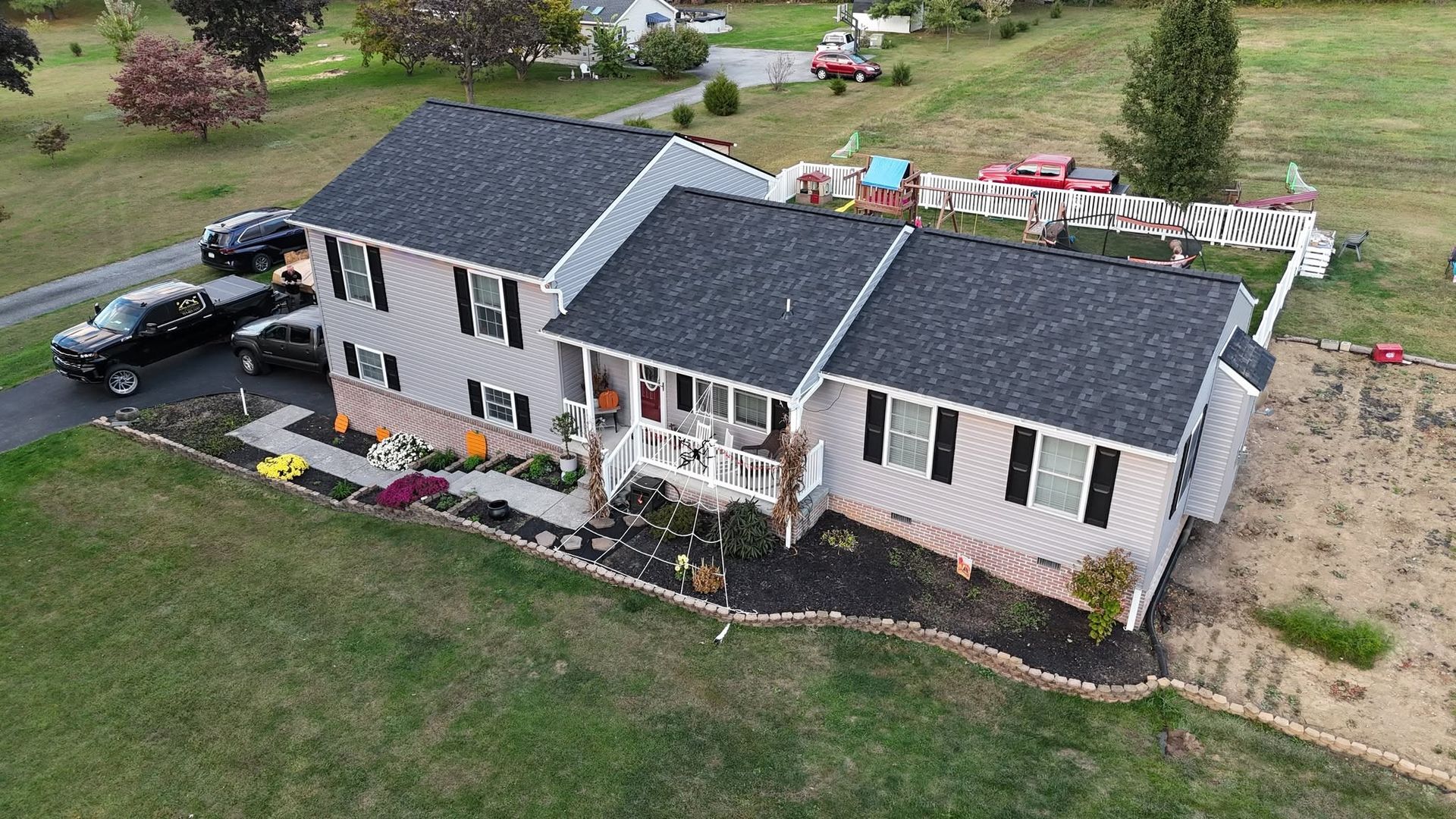Aerial view of a white two-story house with dark roof, front porch, and parked vehicles beside a grassy yard.