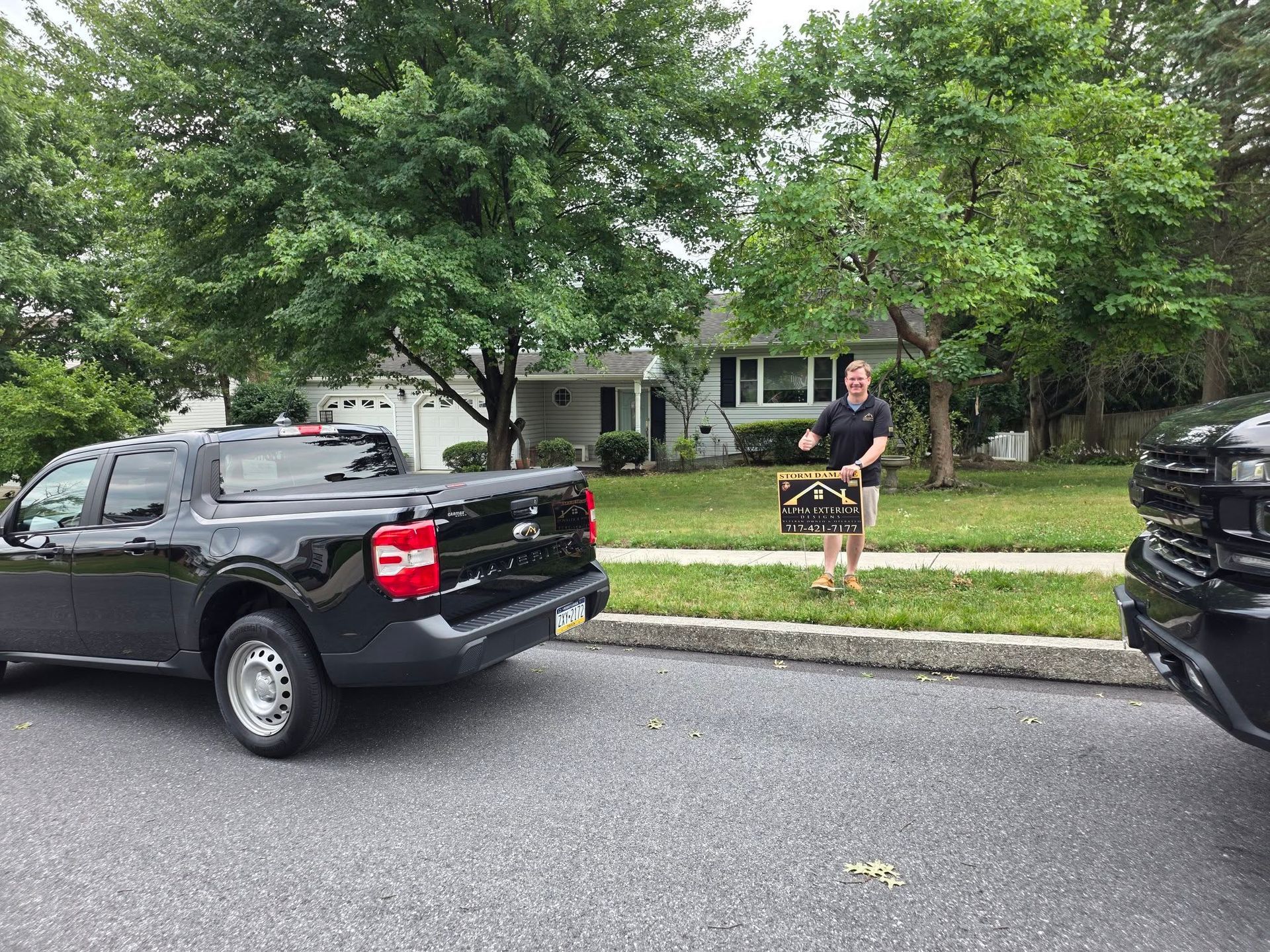 Person standing on a suburban sidewalk between parked pickup trucks, holding a striped bag, with houses and trees behind.