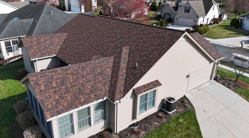 Aerial view of a suburban house with brown shingle roof, beige siding, and a driveway.