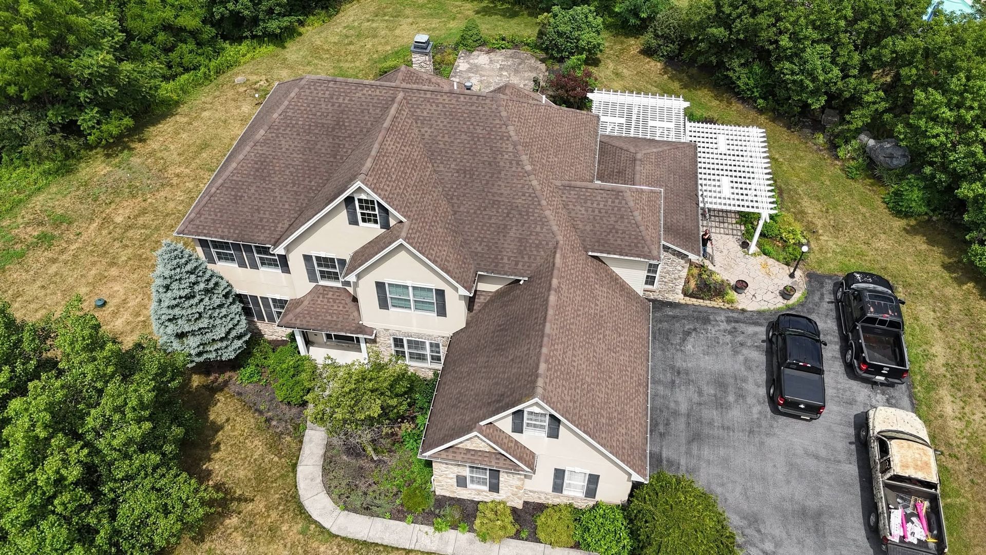 Aerial view of a beige house with a brown roof, driveway, parked cars, and surrounding green trees.