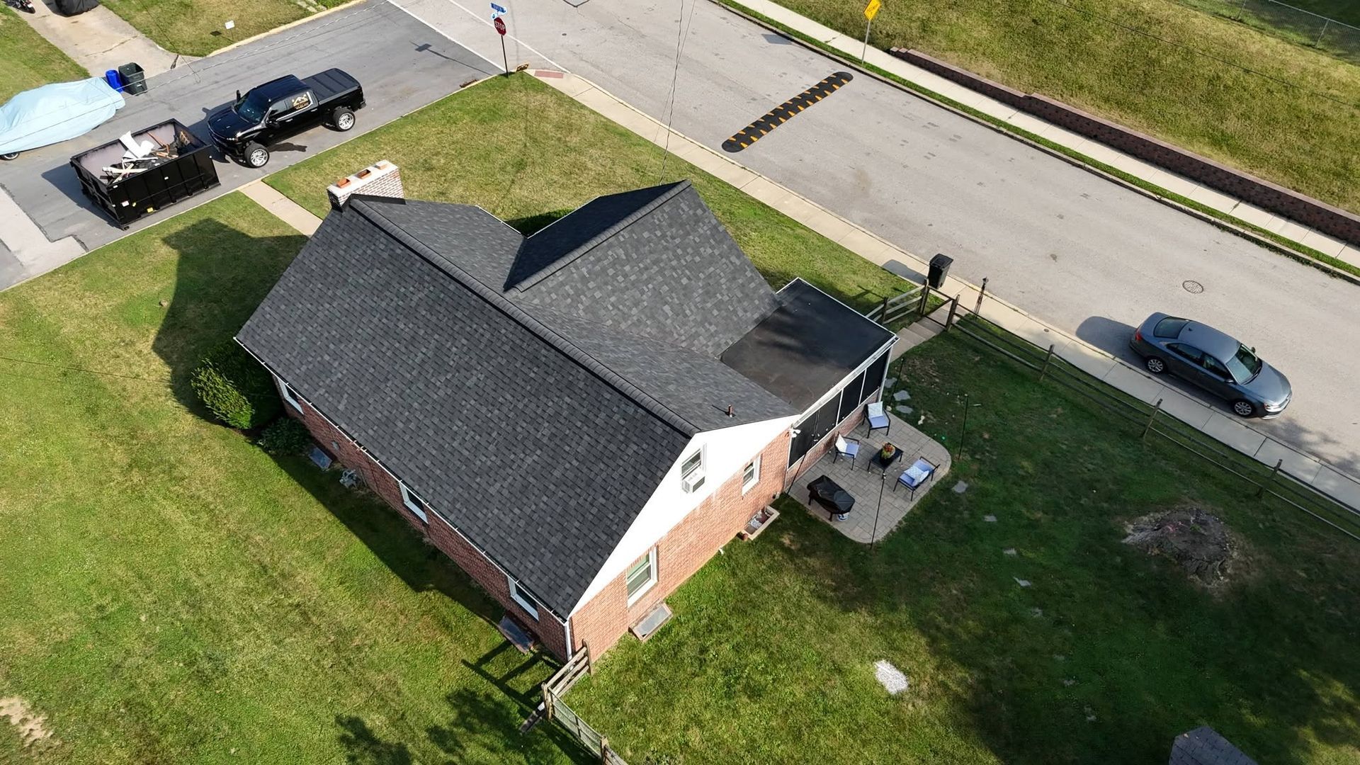 Aerial view of a brick house with a dark roof, lawn, and parked cars along the street.