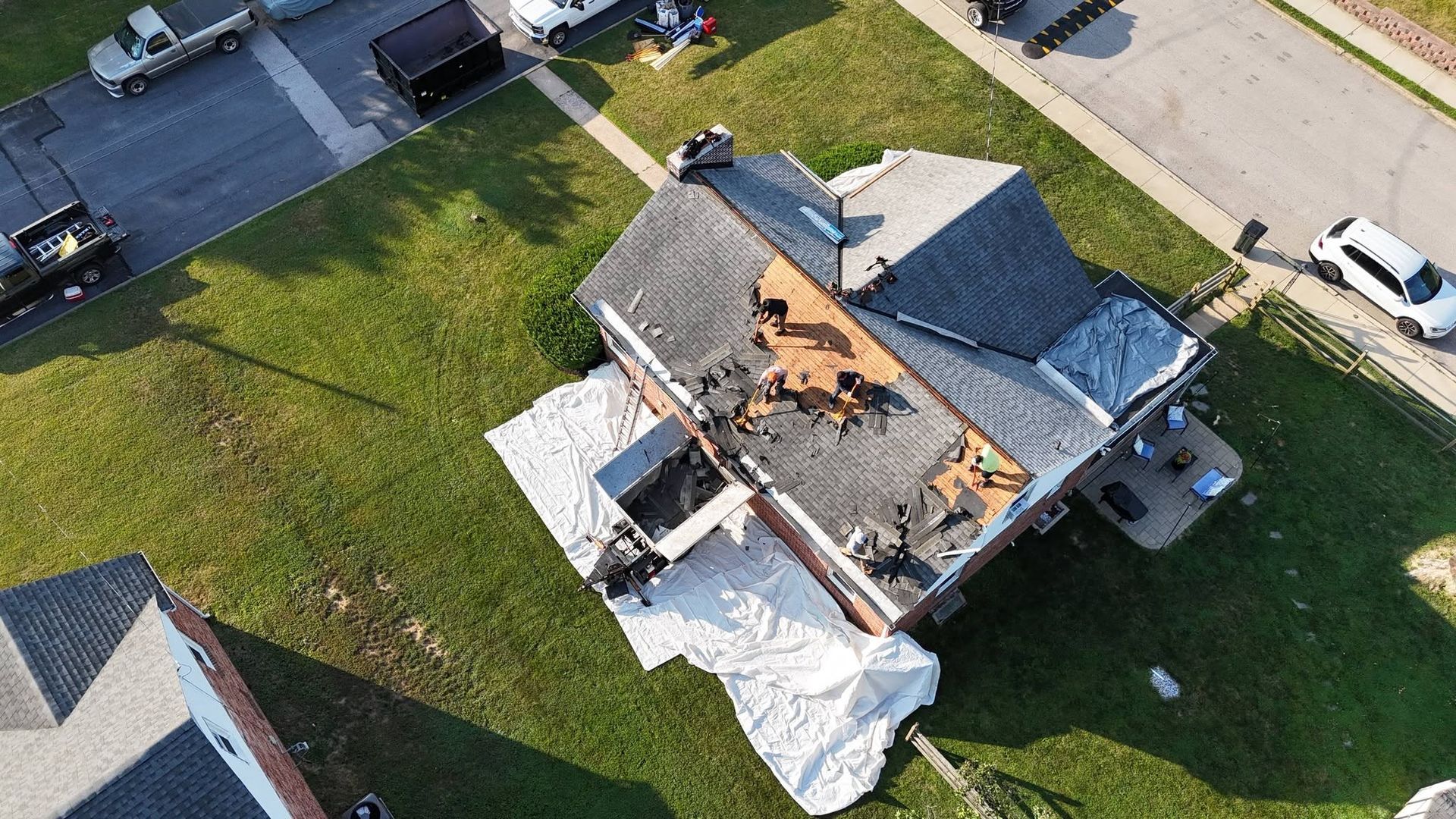 Aerial view of a house with major roof damage and debris, surrounded by grass and nearby parked vehicles.