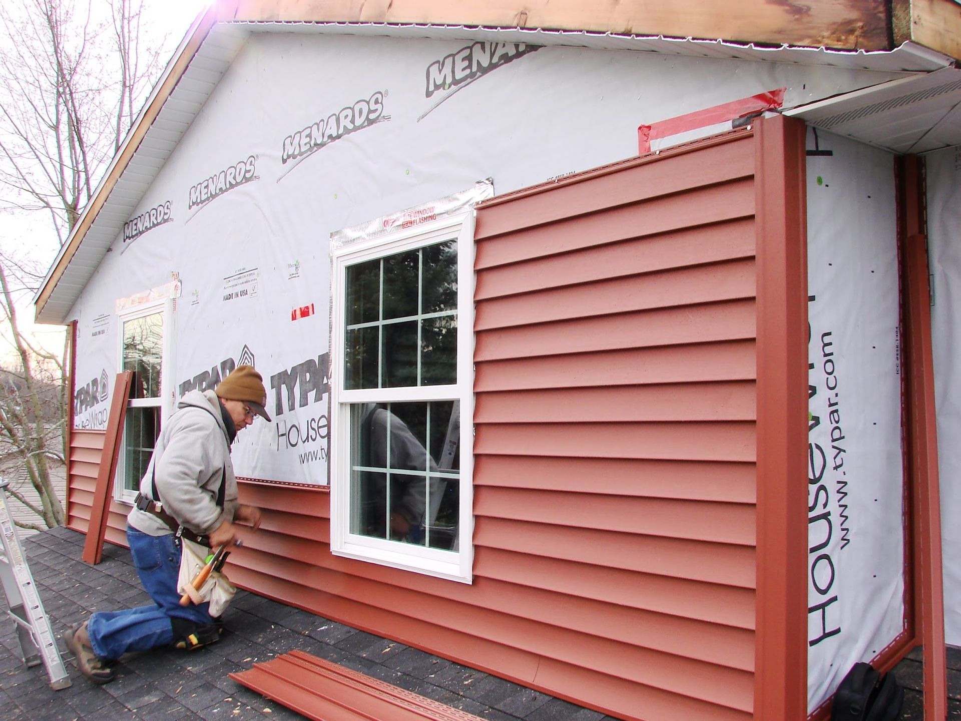 Worker installing red siding on a house exterior with a window and house wrap visible