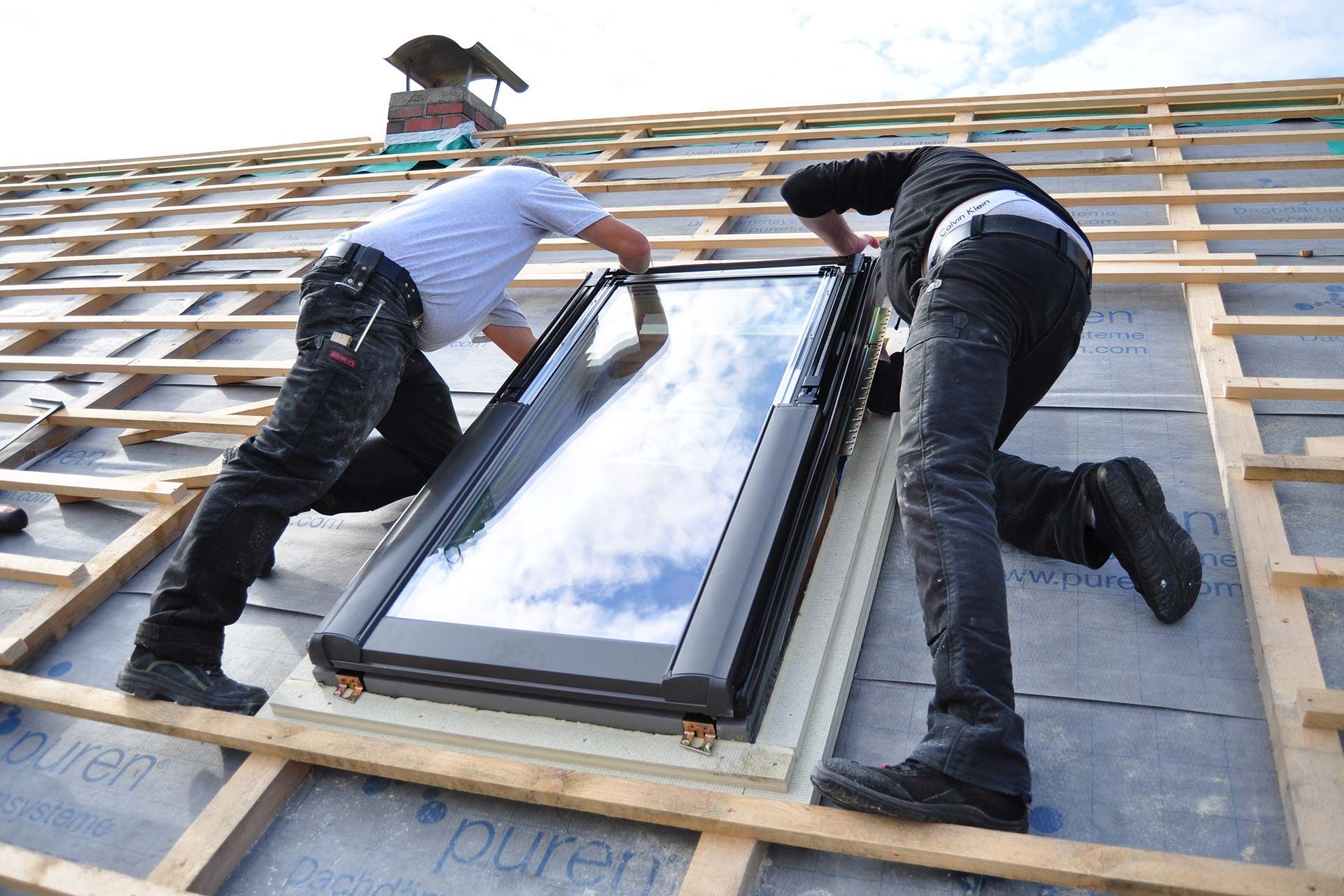 Two workers install a skylight on a roof, framing and securing the window panel.
