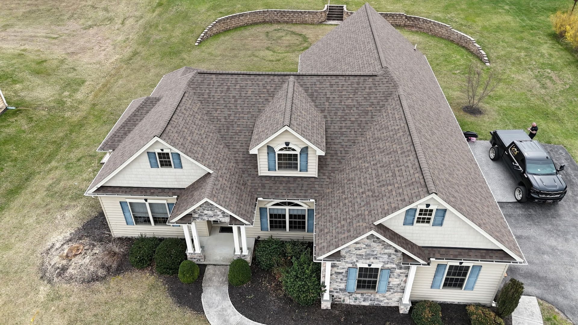 Aerial view of a large beige suburban house with a gray roof and a driveway with a parked black car.