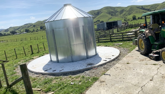 Large metal farm grain bin sitting on a strong agricultural concrete foundation pad.