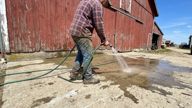 Farmer washing down a durable concrete barnyard paving slab in front of a red barn.