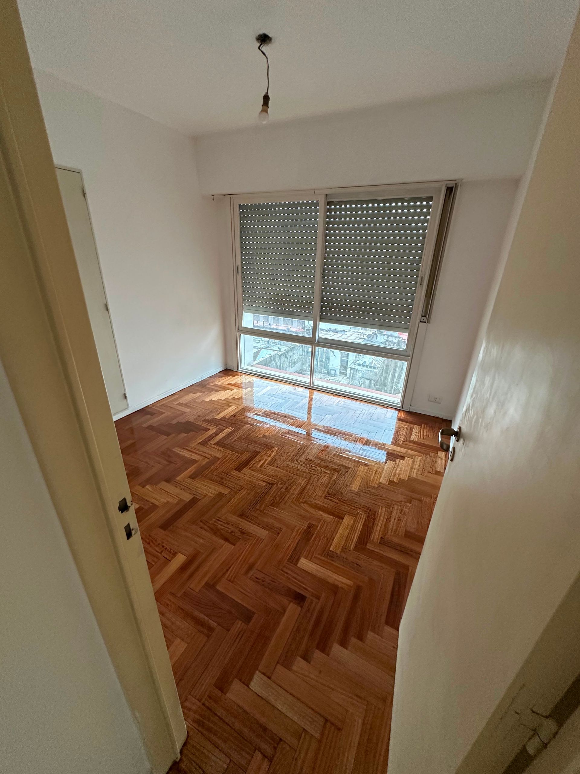 Habitación vacía sin amueblar, con suelo de madera en espiga y luz solar que entra por las persianas cerradas.