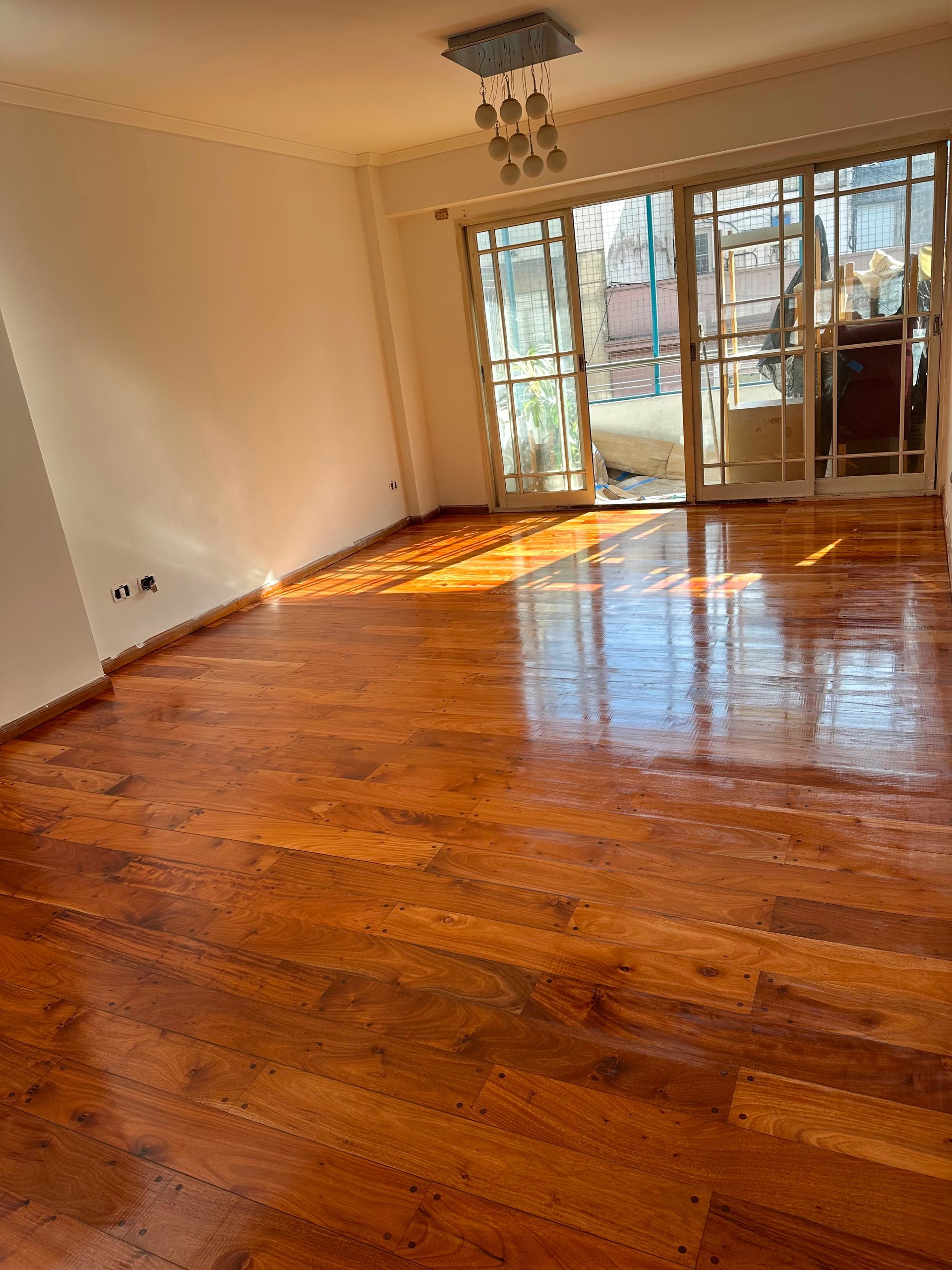 Habitación vacía y luminosa con suelos de madera noble pulida y grandes puertas de cristal que dan a una terraza.