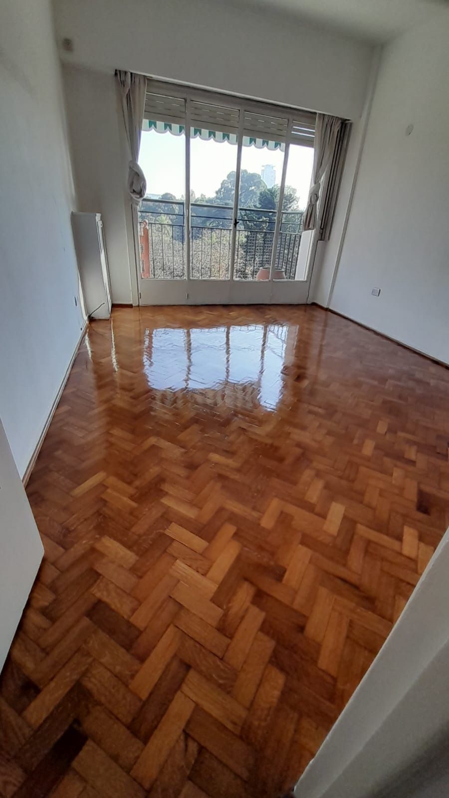Habitación vacía con suelo de madera brillante en espiga y grandes puertas correderas de cristal que dan a un balcón.