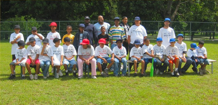 Group of people posing outdoors on a grassy field, seated and standing in front of a fence.