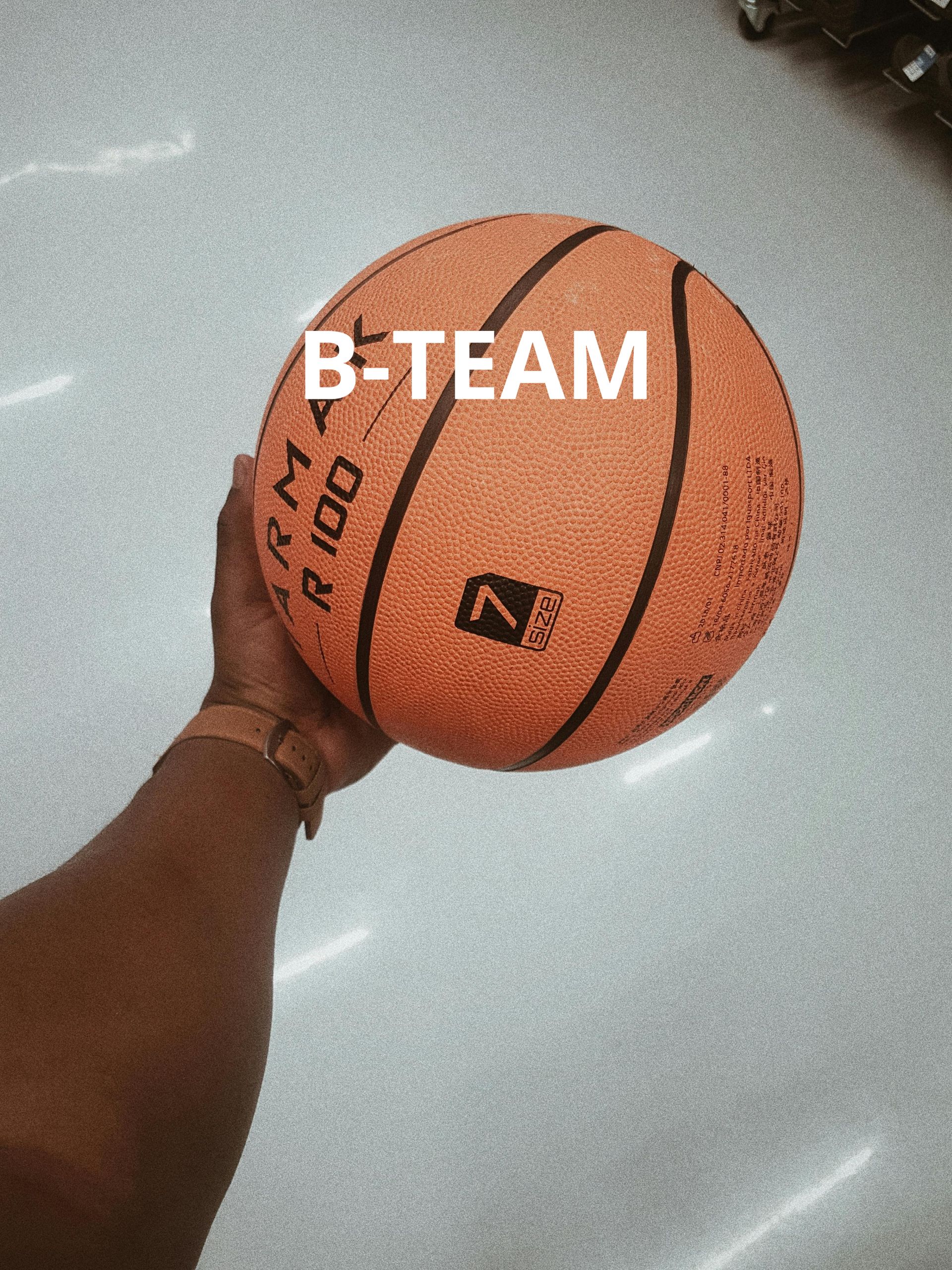 Hand holding an orange basketball with “B-TEAM” text on a gray floor