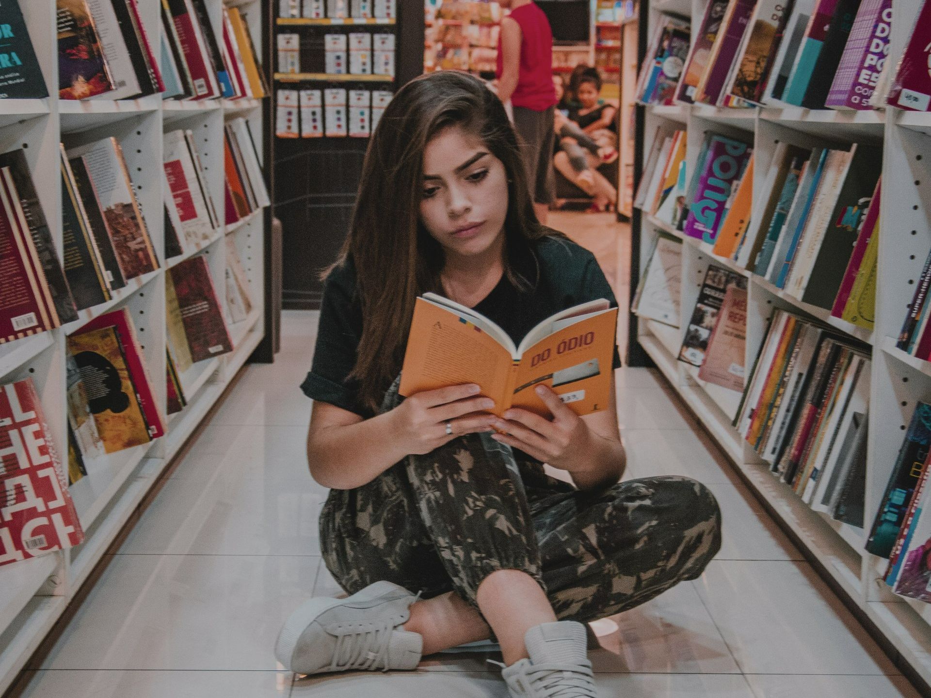 Young woman sits on a bookstore floor reading an orange book between tall shelves.