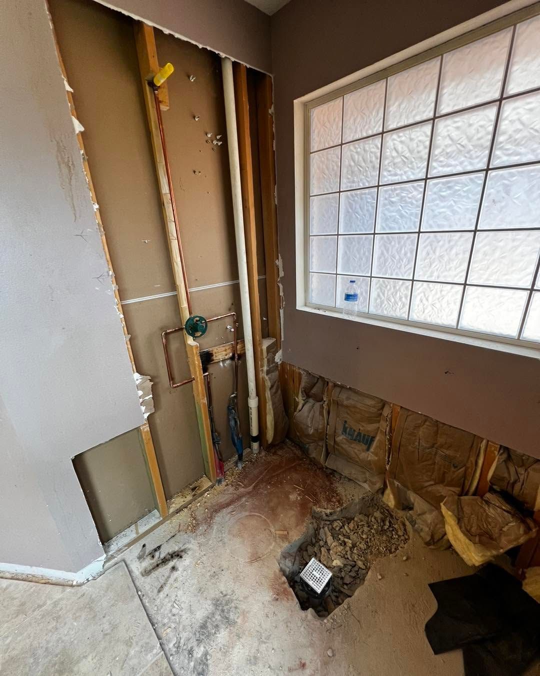 Bathroom under renovation with exposed pipes, torn drywall, and debris by a window.
