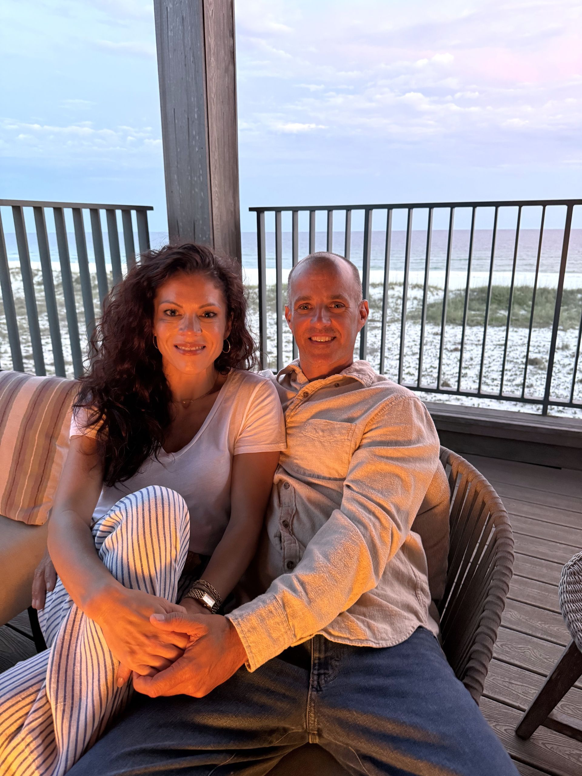 Couple sitting together on a balcony sofa at sunset, holding hands and smiling, with ocean in the background