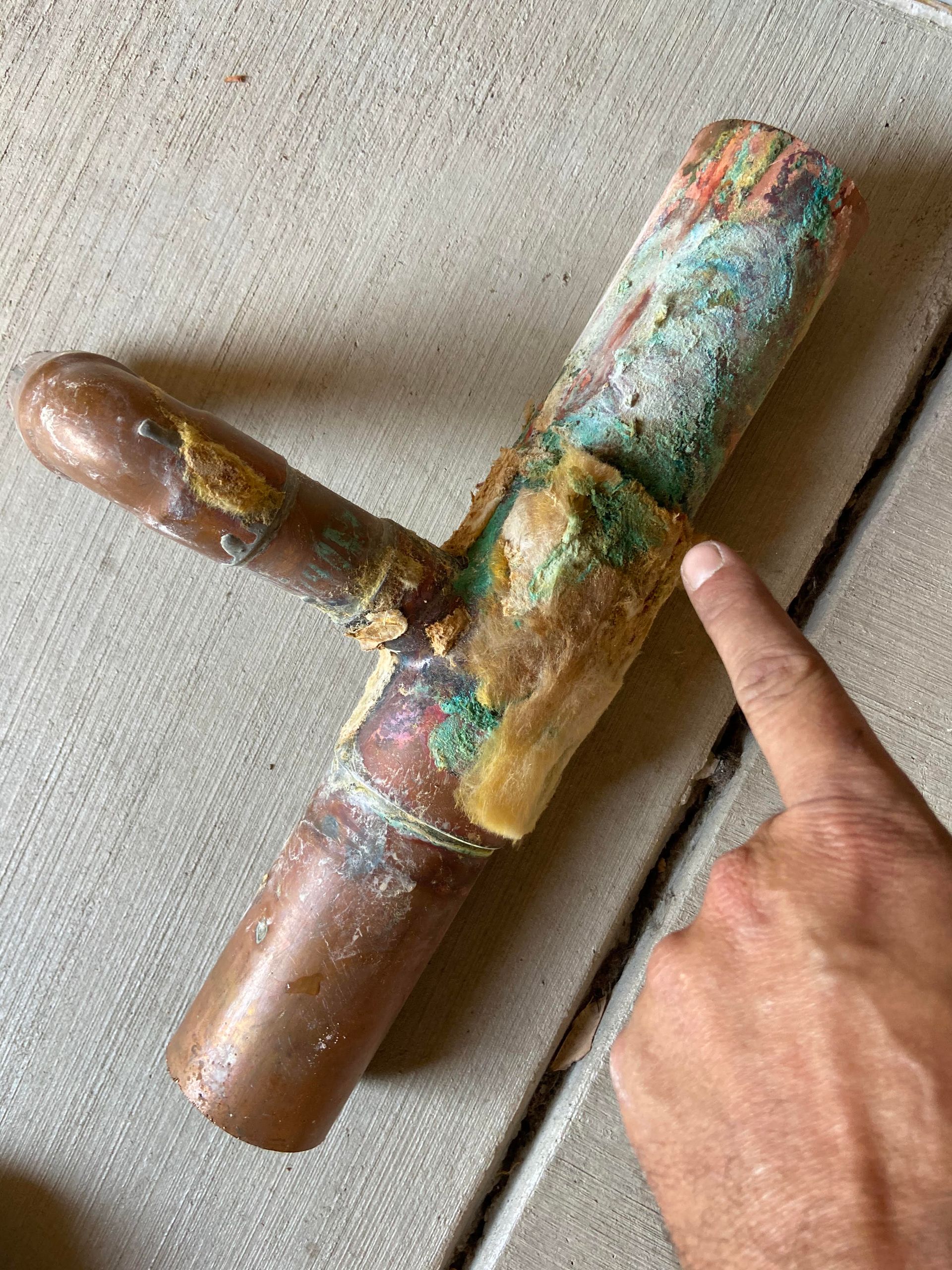 Rusty copper pipe fitting with a T-shape, held by a hand against a concrete floor.