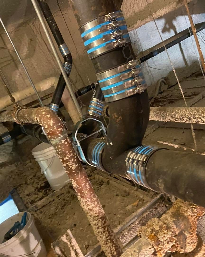Multiple insulated black pipes with blue bands in a wet basement or crawlspace, beside a white bucket and rusted pipe
