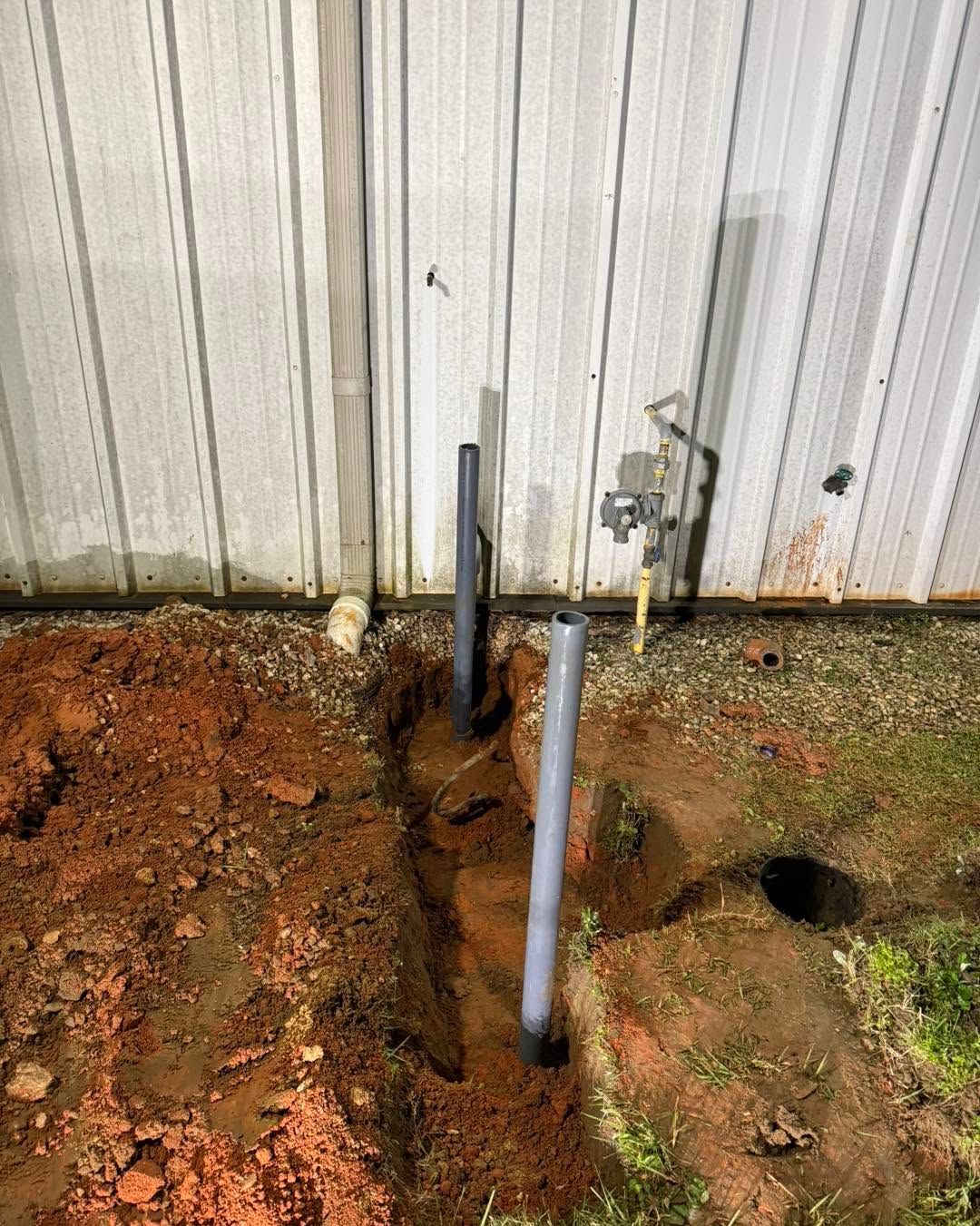 Exposed pipes and a gas meter beside a corrugated metal wall, with dirt dug out around them.