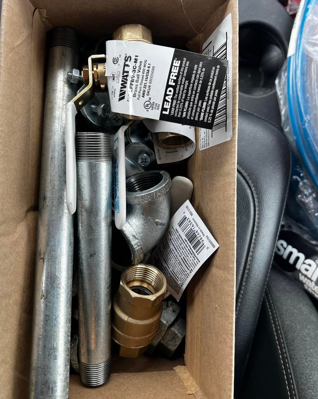 Box of assorted metal pipe fittings and connectors, mostly silver and brass, with product tags.