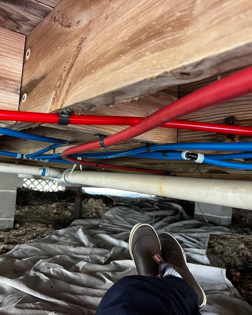 Crawlspace with red and blue pipes under floor joists; a personβs shoe visible on plastic sheeting