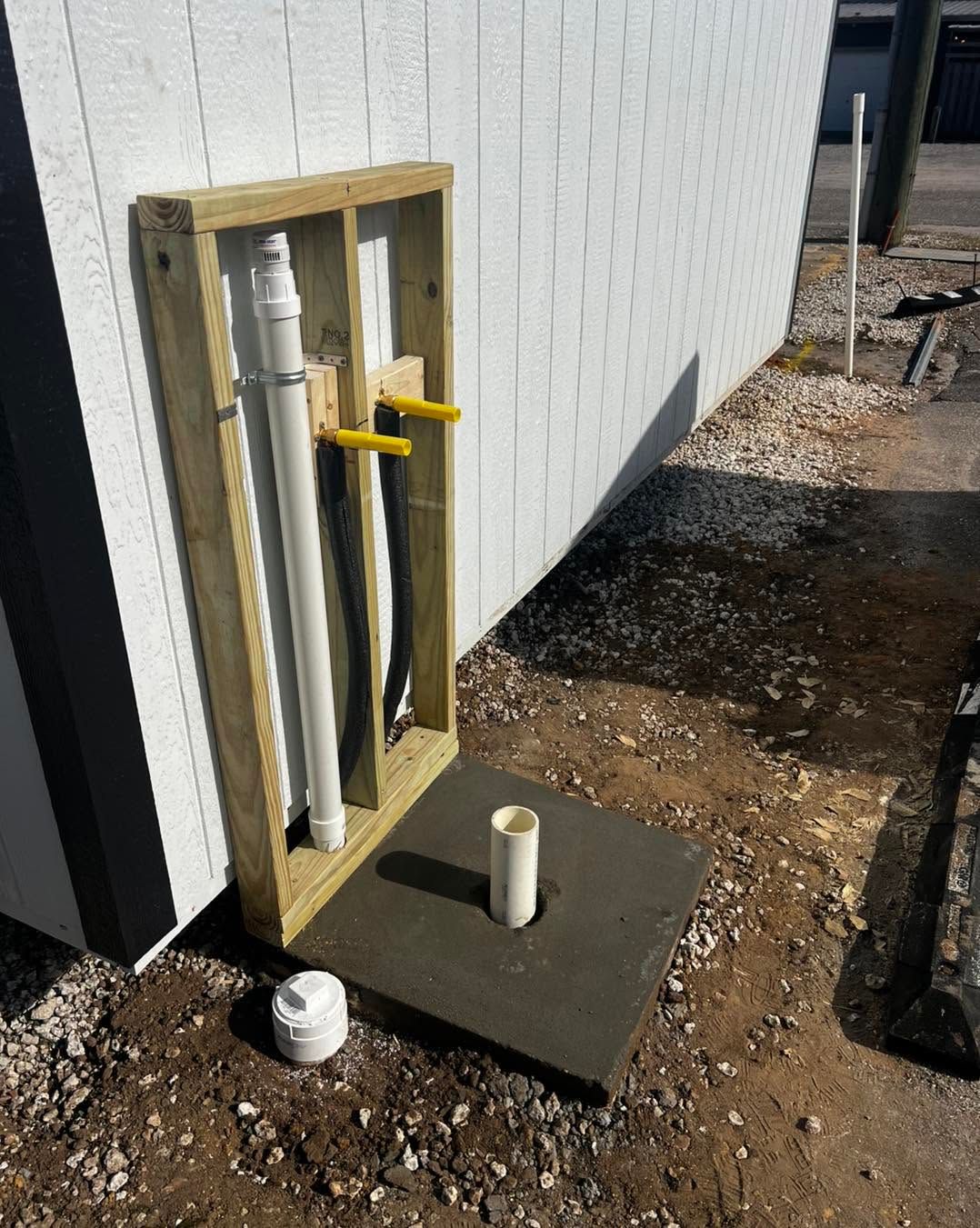 Exposed outdoor plumbing pipes beside a white building wall, with gravel ground and a small concrete pad.