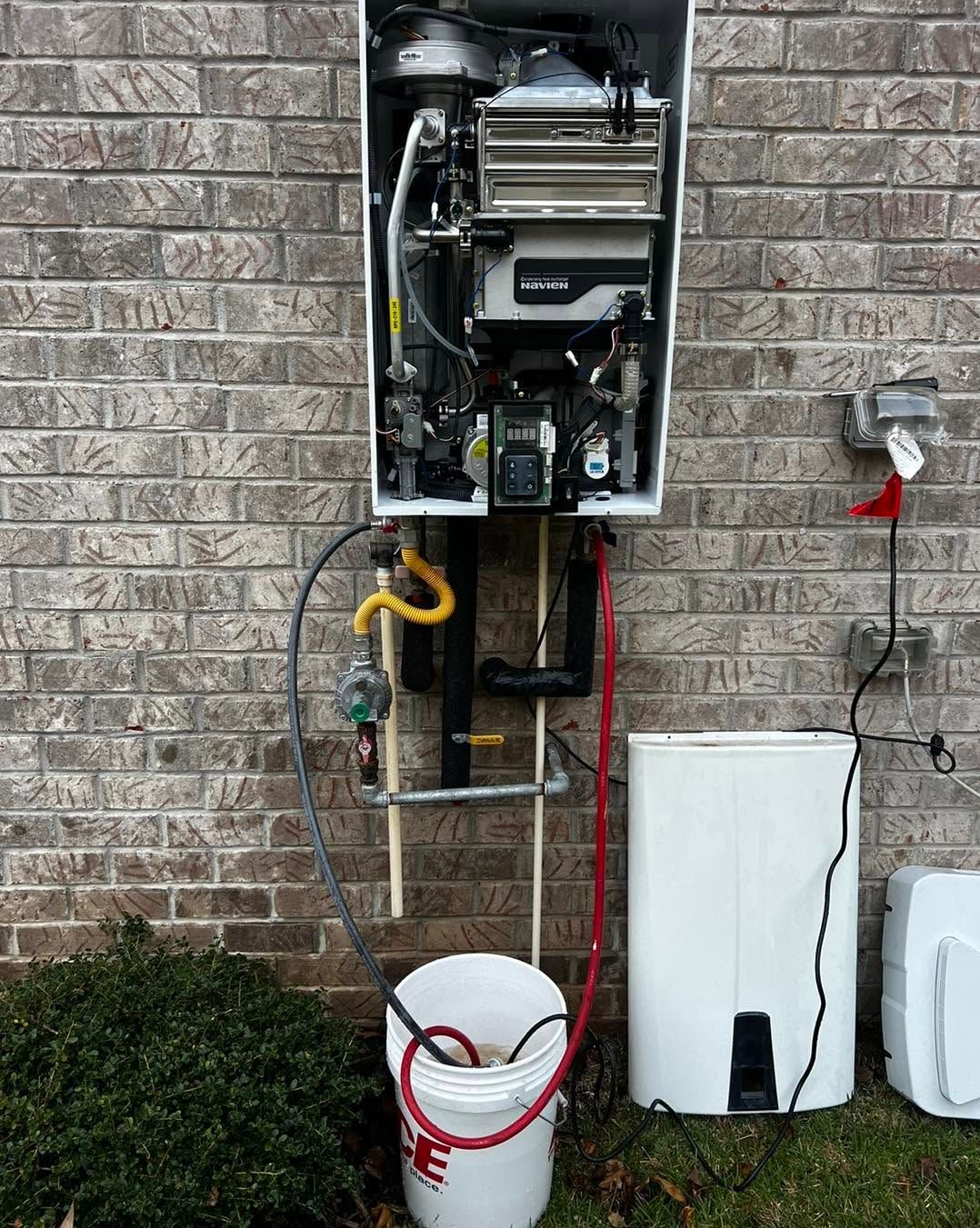 Open electrical meter and utility lines mounted on a brick wall, with hoses and a bucket below.