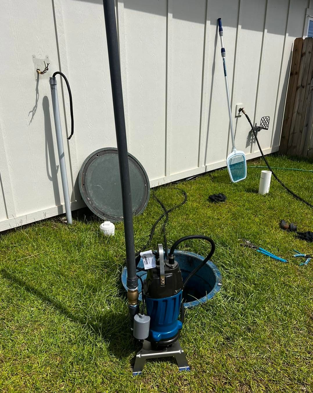 Blue water pump on grass beside a white fence, with hoses and yard tools nearby.