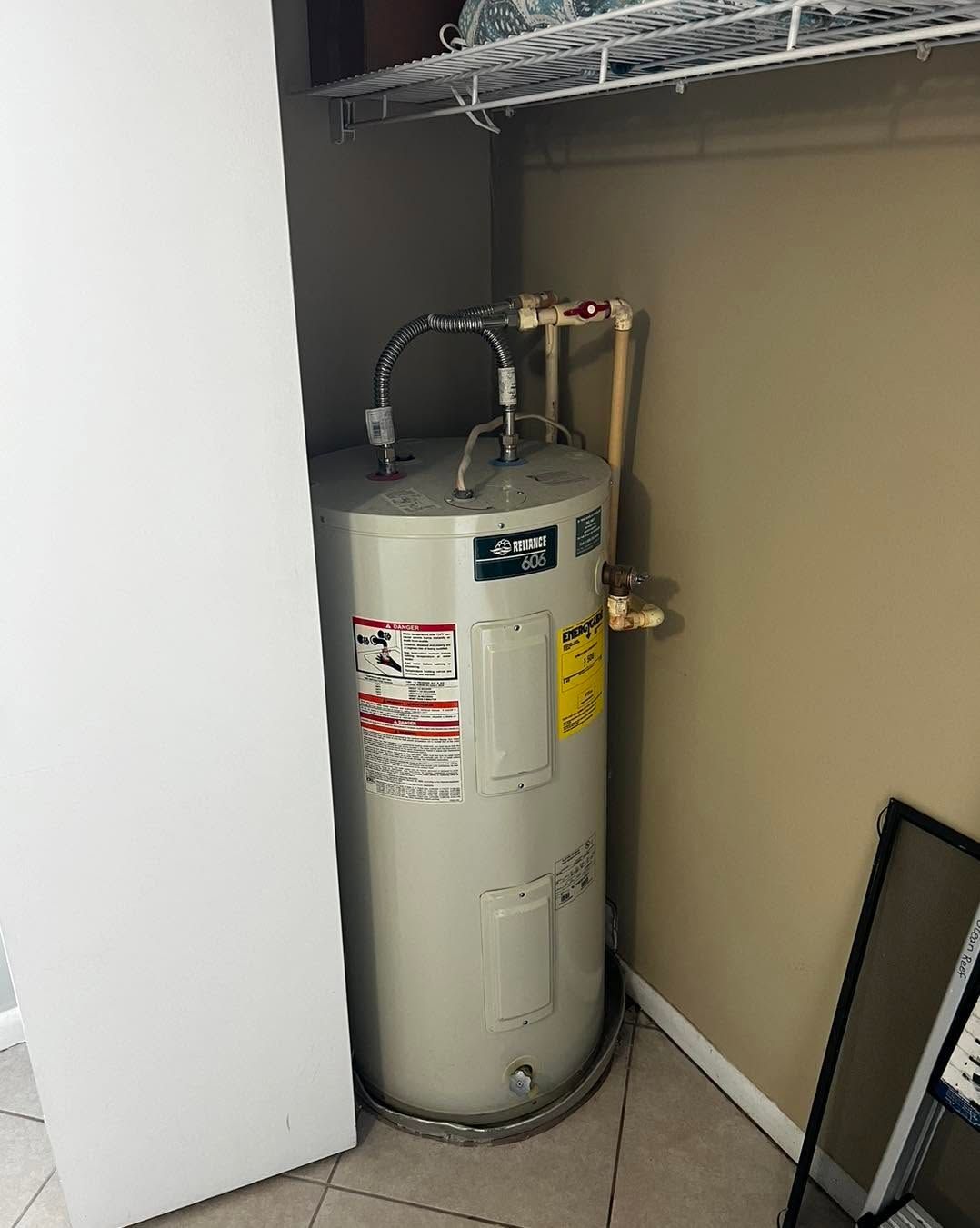 Water heater in a closet with exposed pipes and shelving above.