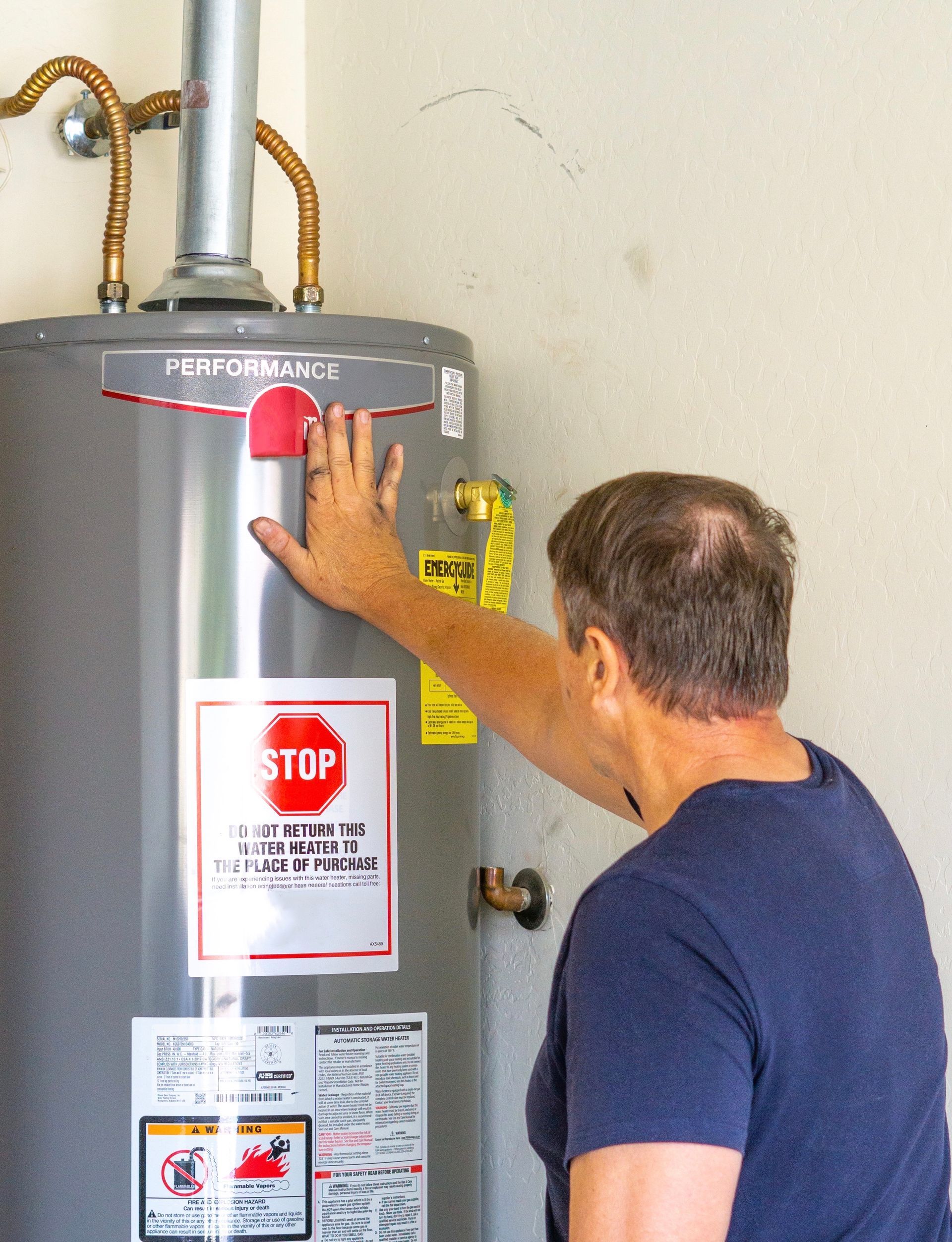 Man touching a tall, silver water heater. He is in front of a light-colored wall.