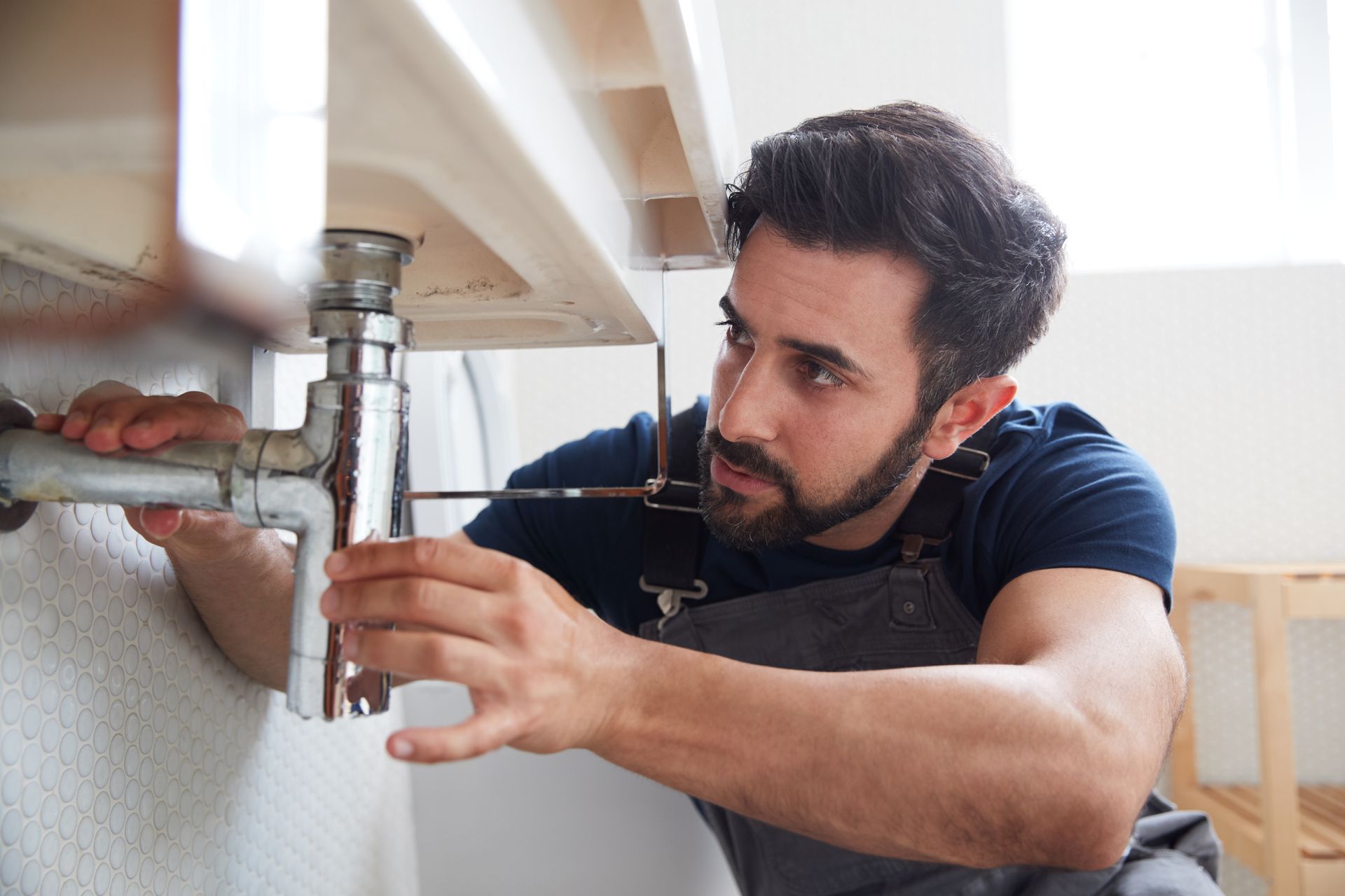 Plumber working under a sink, using a tool. He is focused on the plumbing, in a bathroom.