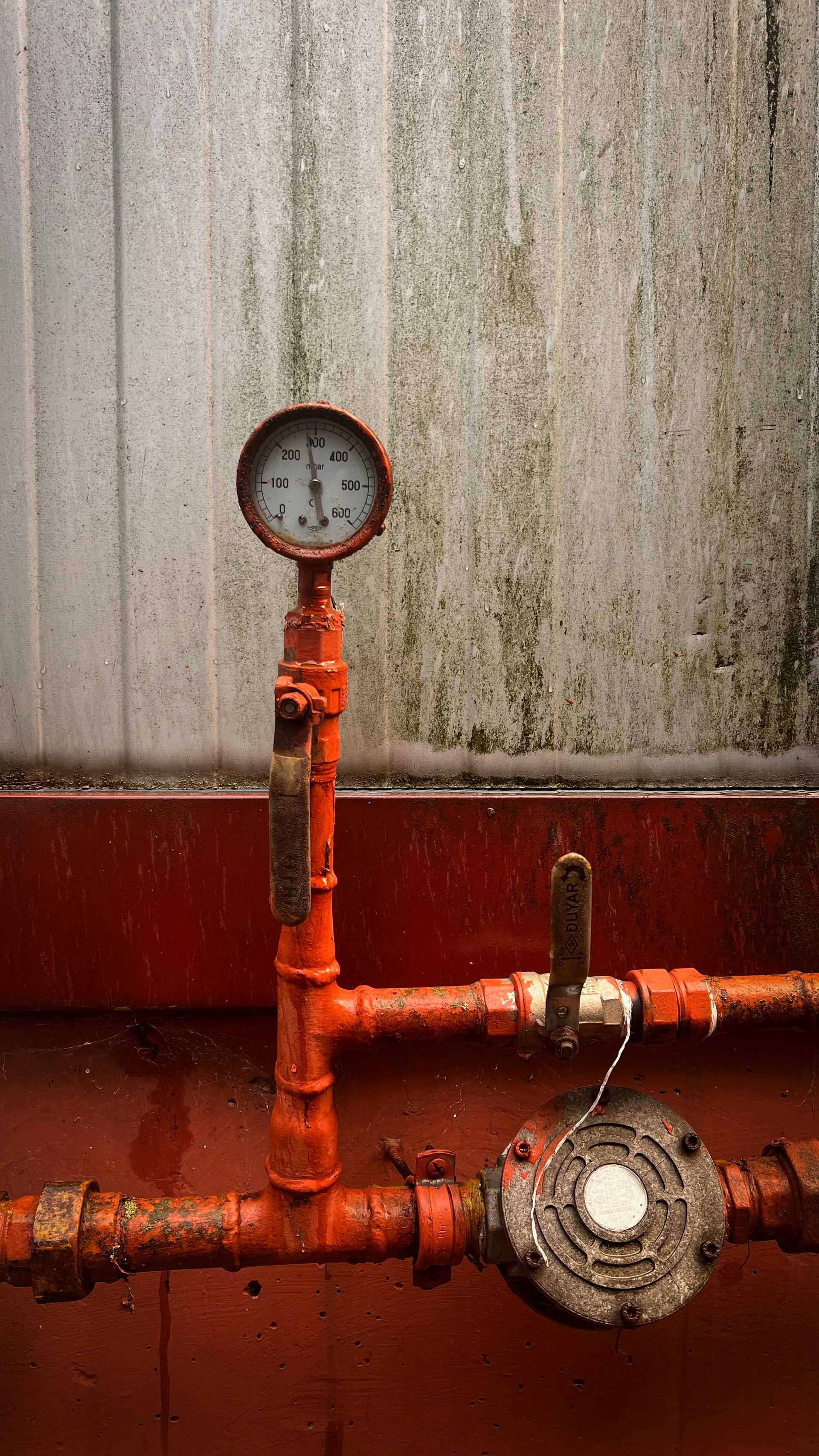 Rusty red industrial pipes with a pressure gauge against a stained wall, showing a valve and fittings