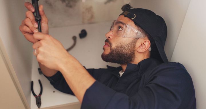 Man working on plumbing under a sink. He wears safety glasses, a cap, and holds a wrench. Smoke present.