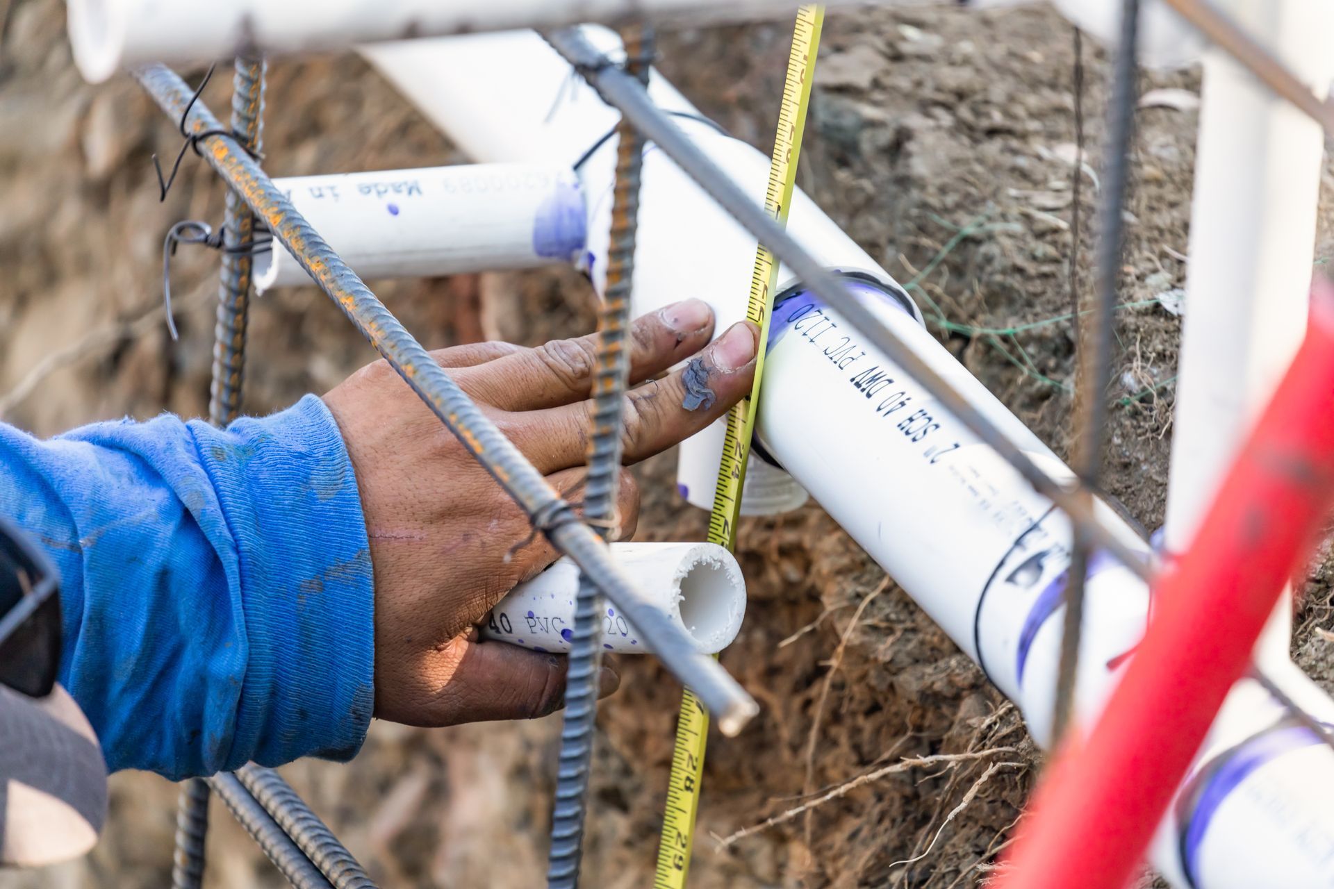 Hand measuring a PVC pipe with a measuring tape amidst rebar and other pipes on a construction site.