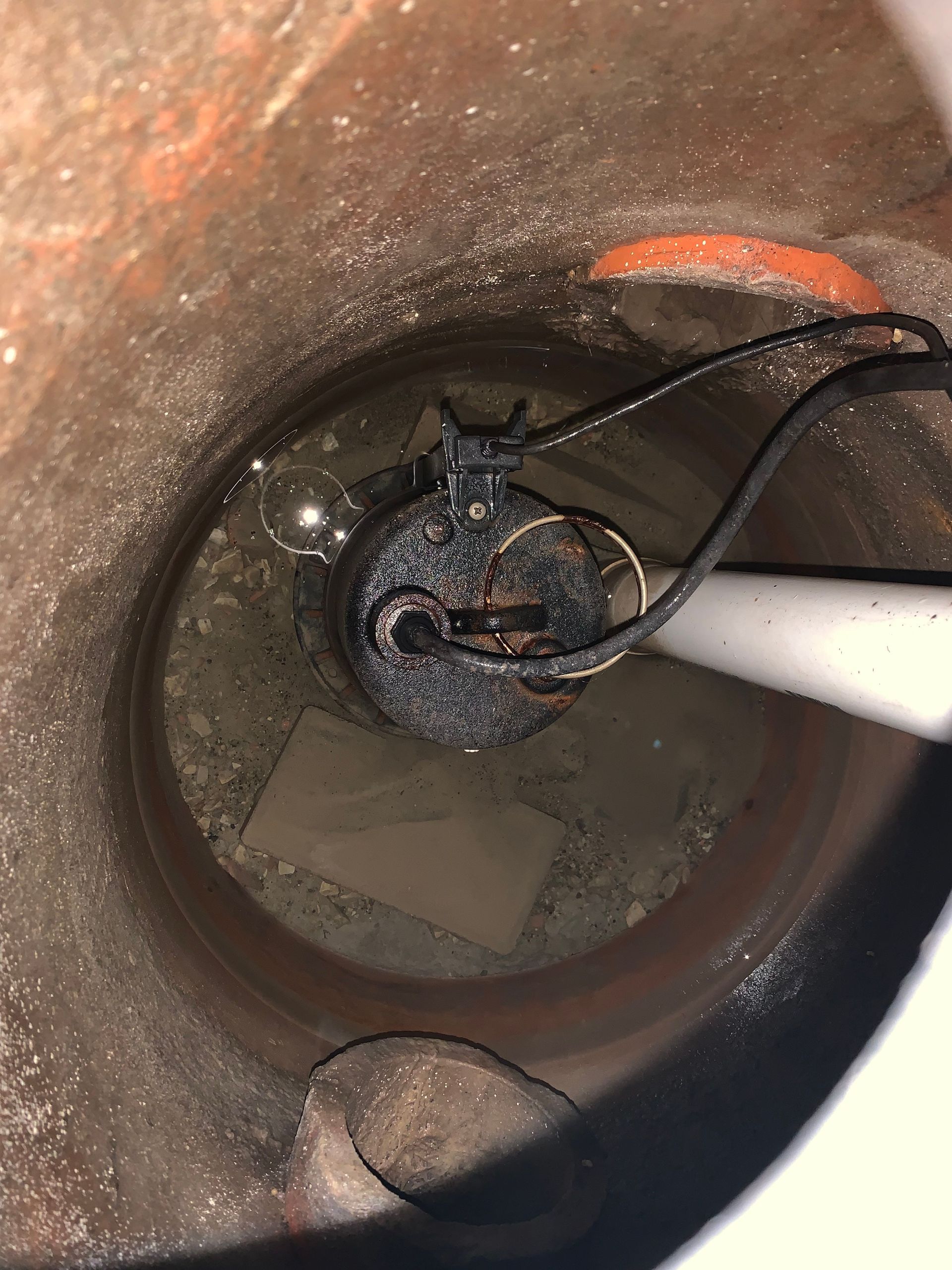 Looking down into a sump pit with a submerged pump and debris. Black wires and a white pipe are visible.