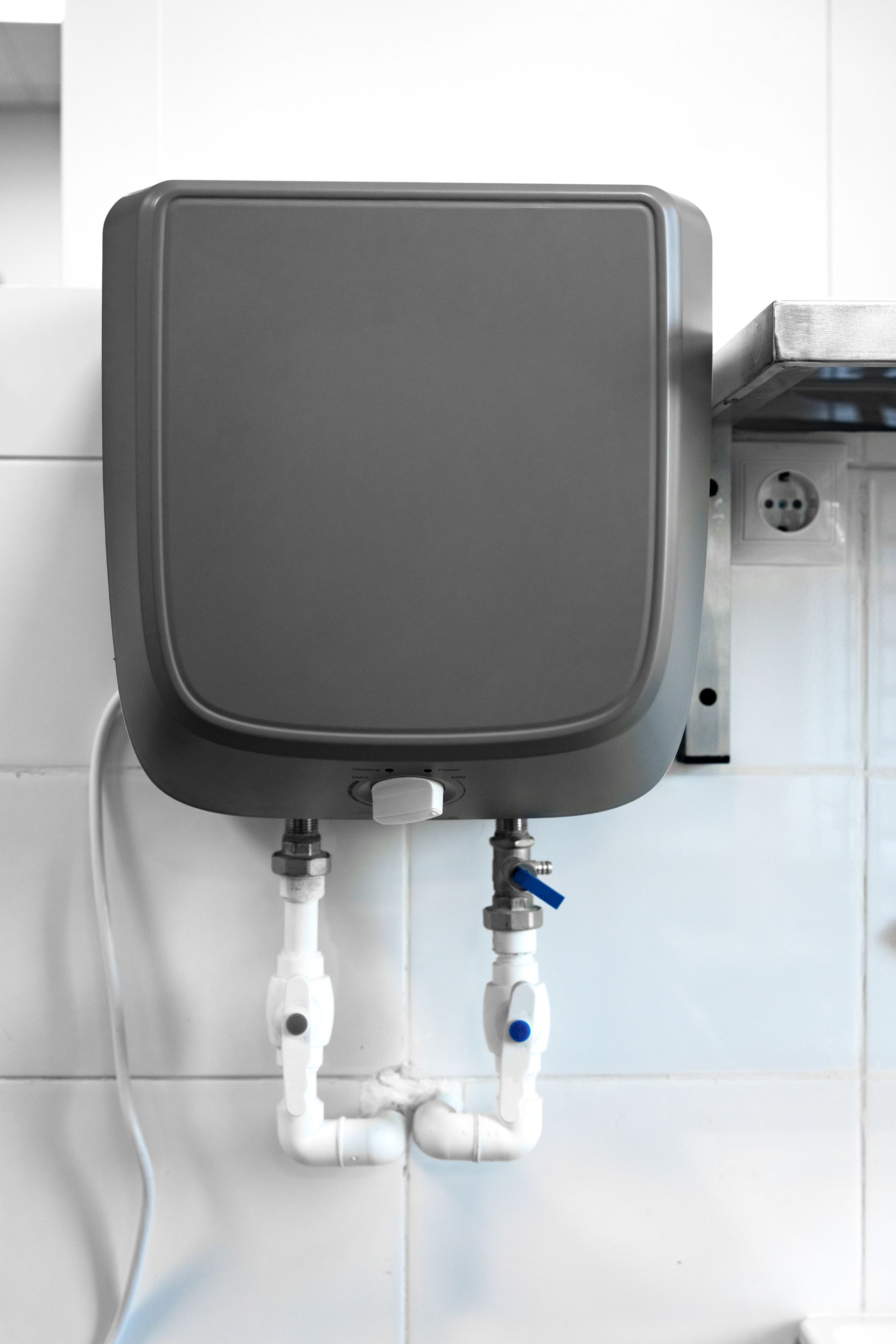 Gray electric water heater mounted on white tiled wall. White pipes connect to the unit.