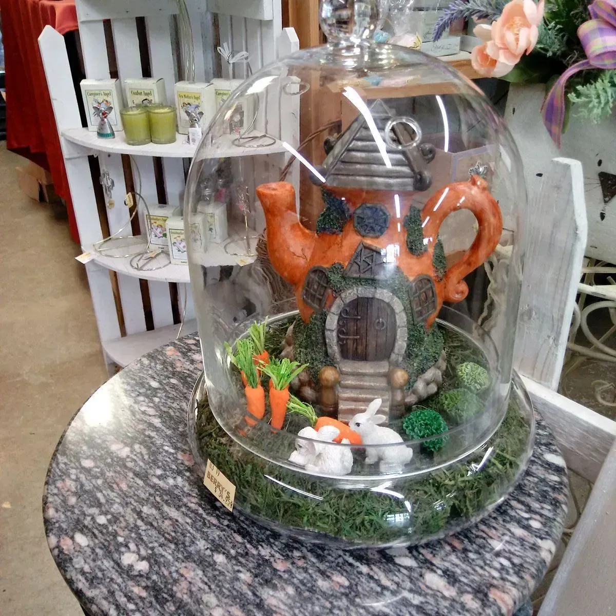 A whimsical miniature fairy garden inside a glass dome with a teapot house and small bunny and carrots.